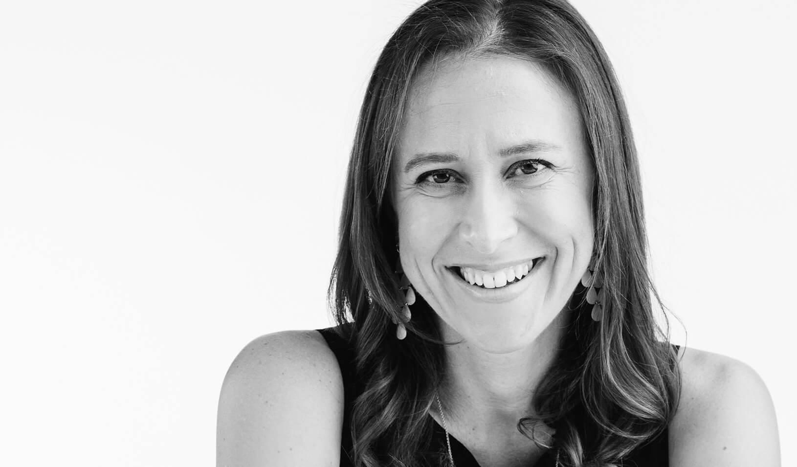 Black and white portrait of Anne Wojcicki smiling at the camera on a white background. Courtesy of Anne Wojcicki