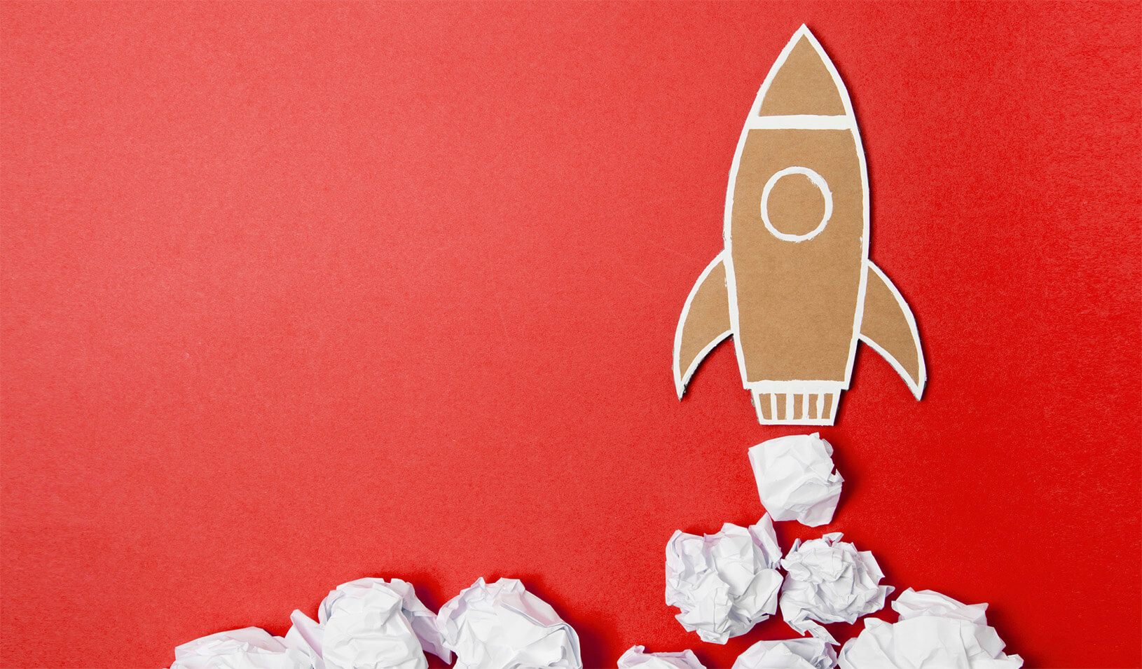 A rocket ship cut out of cardboard paper flies up over a red background, with smoke made of clouds under the rocket ship. iStock/mattjeacock