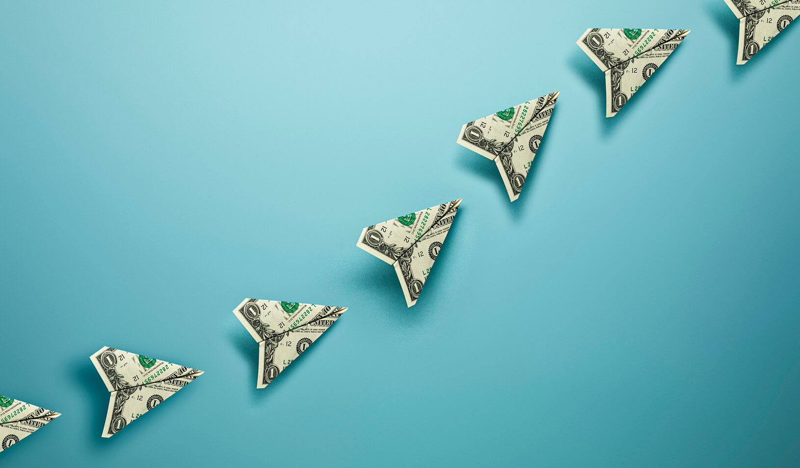 Dollar bills folded into paper planes ascending upward across a blue background. iStock/skodonnell