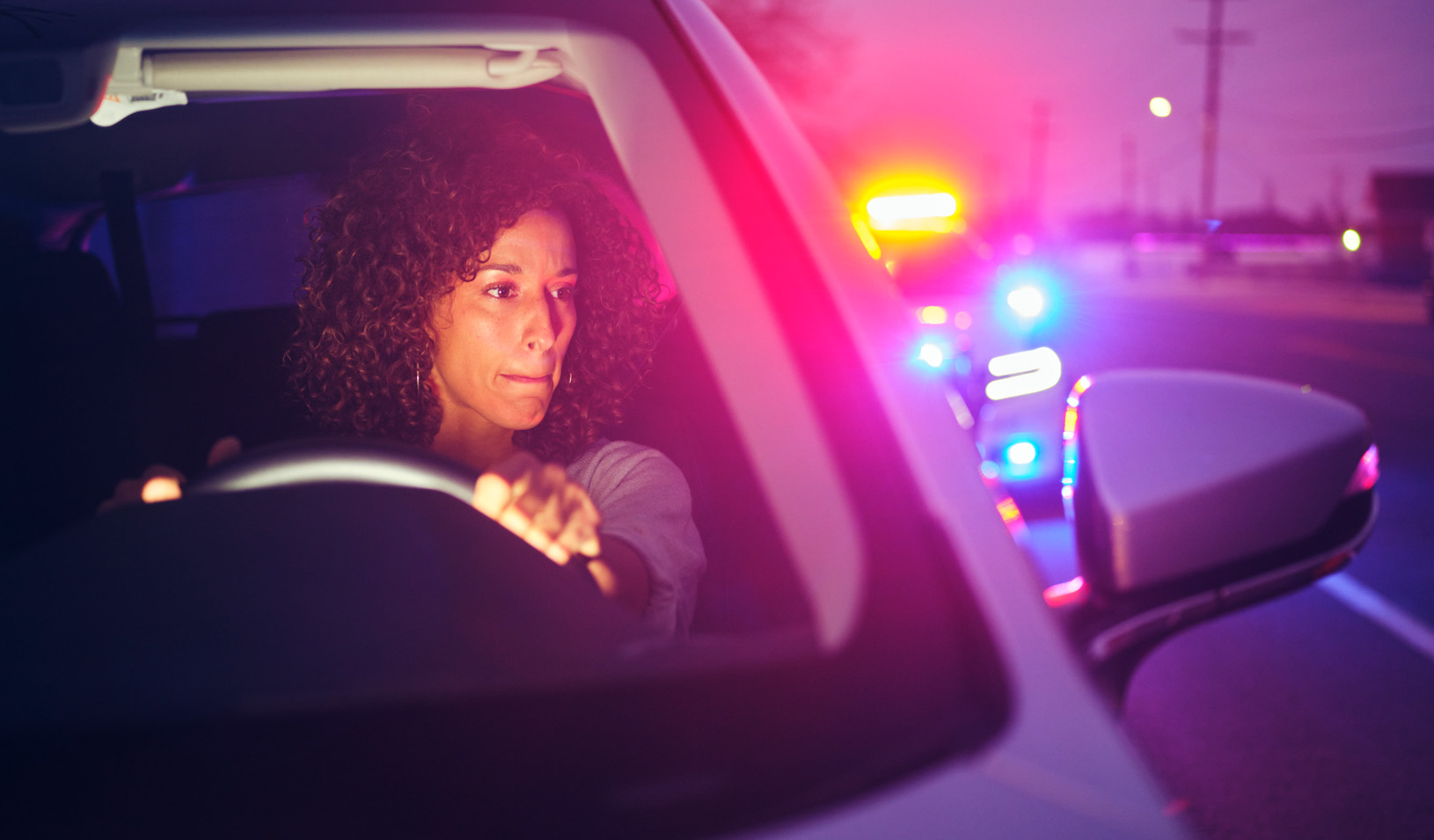 A photo of a Black woman in a car, looking nervously back at the flashing lights of the police car that has just pulled her over. Credit: iStock/RichLegg