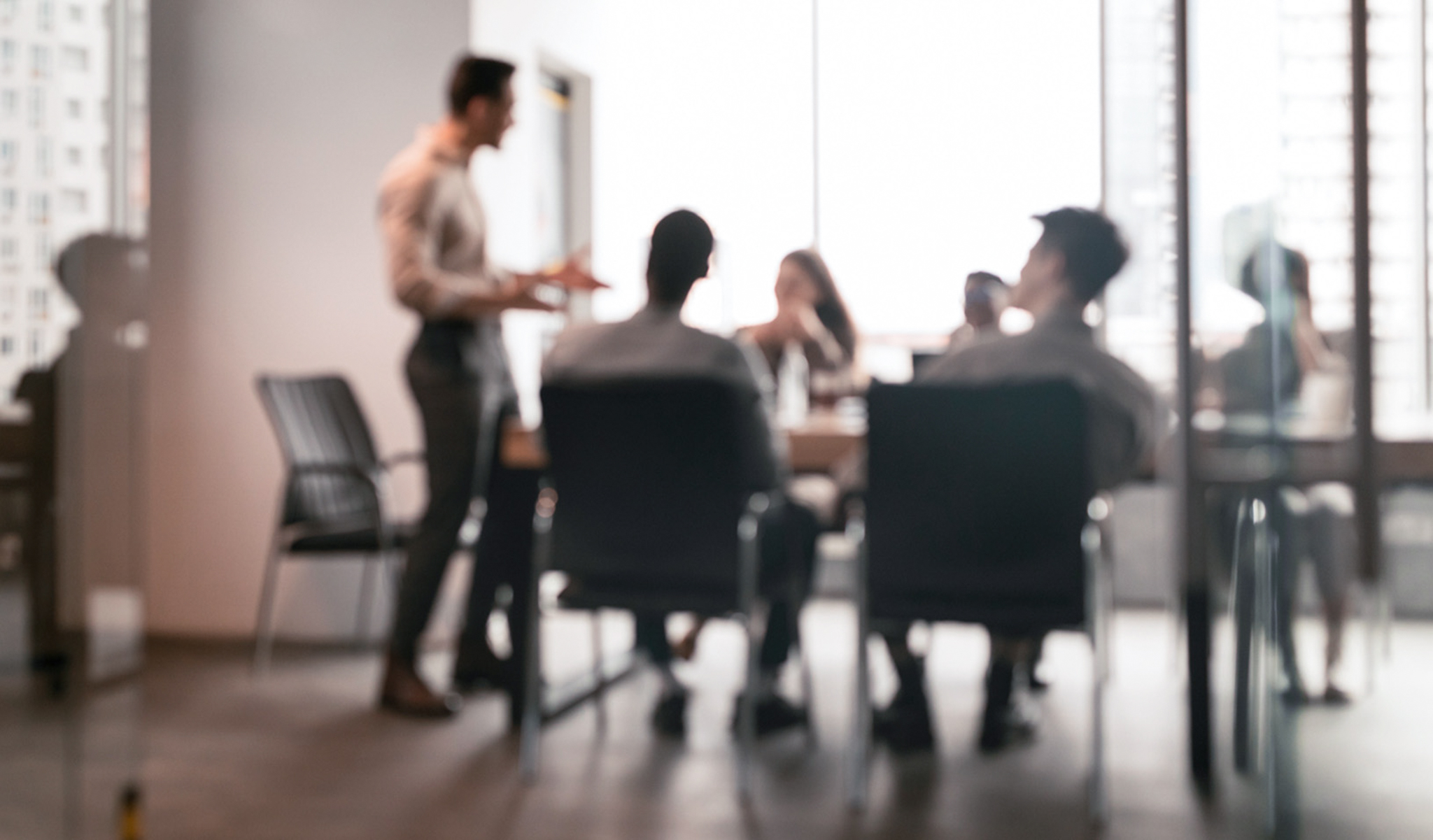 A blurry photo of a boardroom with figures standing and sitting illuminated by a bright window.