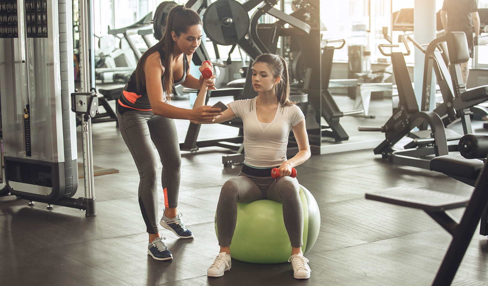 Two women in a gym, one is a trainer looking at her phone, and the other is sitting on an exercise ball.