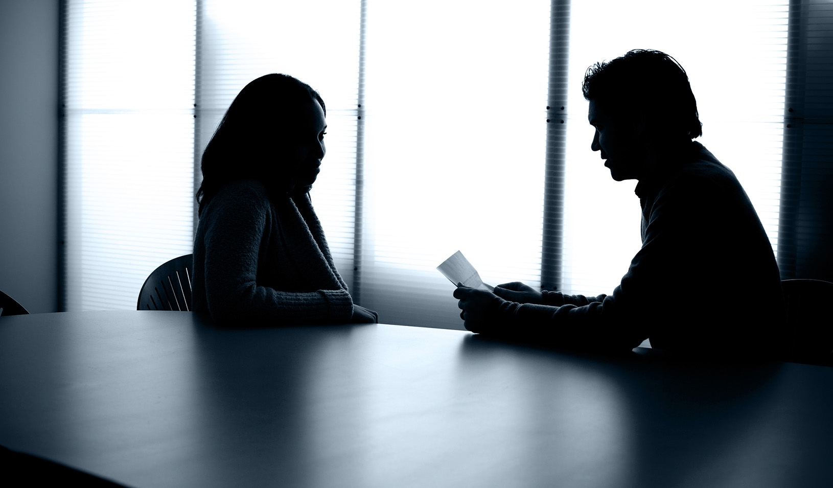 Two people sitting at a conference table in a backlit room, one of them is holding up a piece of paper in front of the other.