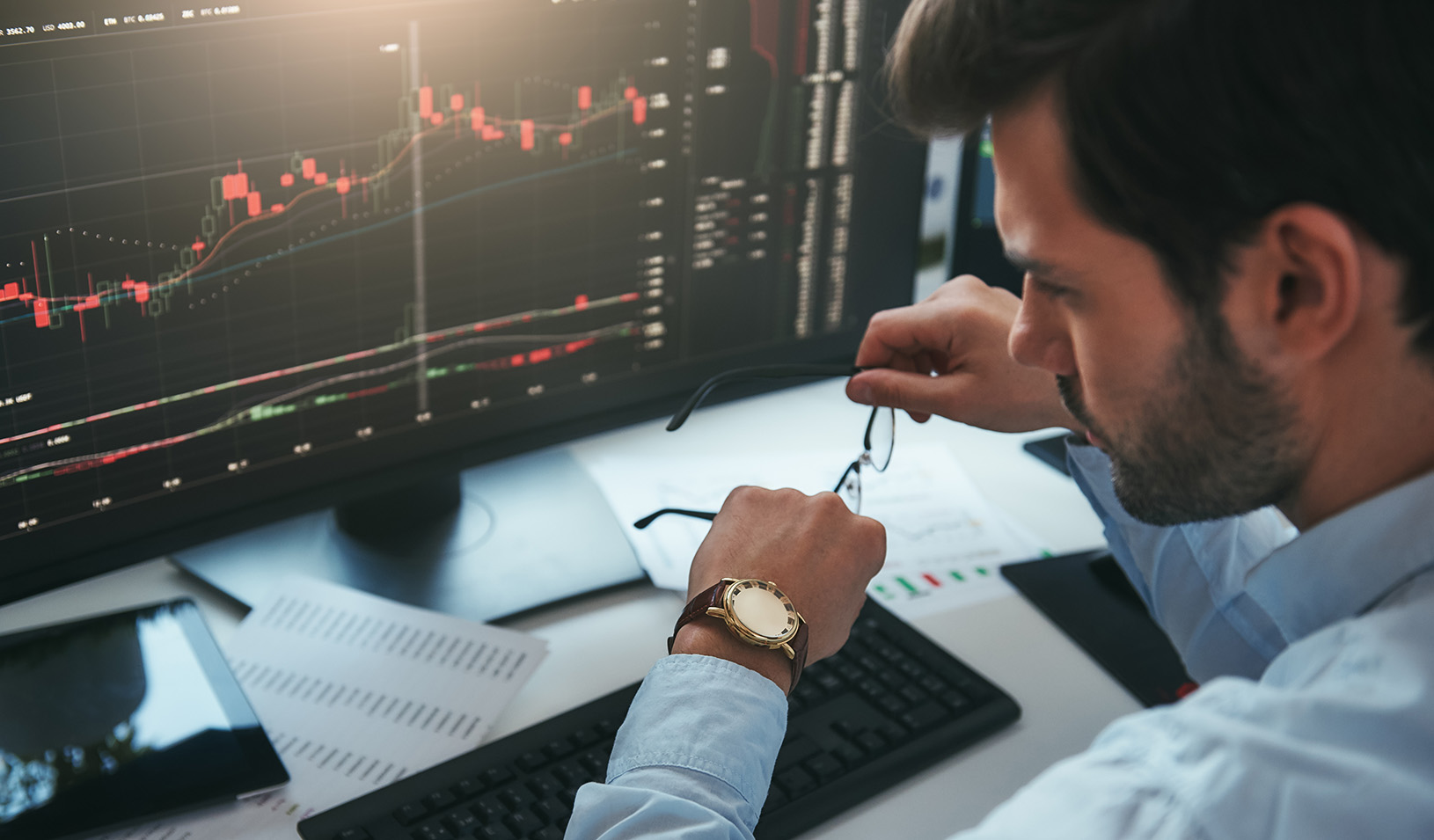 Stock trader looking at watch on hand while working with data and charts on computer screens in an office.