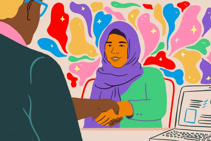 Alt text: A woman in a hijab sitting across a desk from a man in a suit, shaking his hand and smiling. | Credit: Sol Cotti