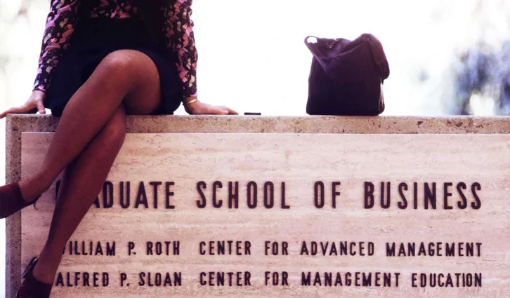 Older photo of a women sitting atop a sign for the Graduate School of Business. Credit:  TBD