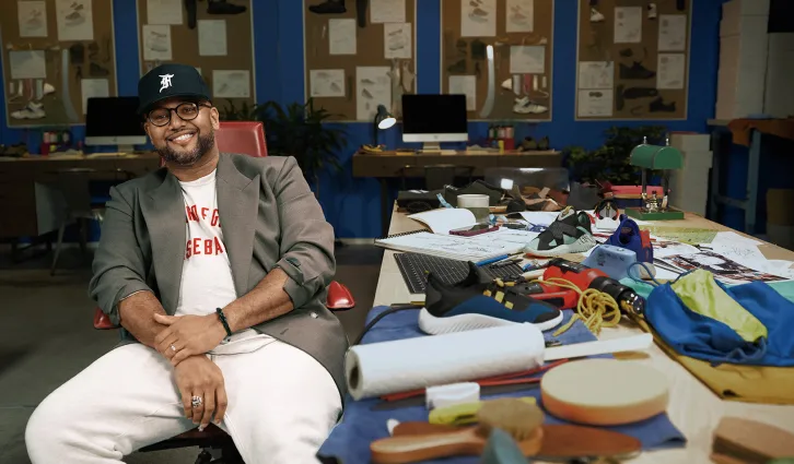 Jason Mayden smiling, sitting in his workshop at his desk, which is covered in materials, papers, and tools.